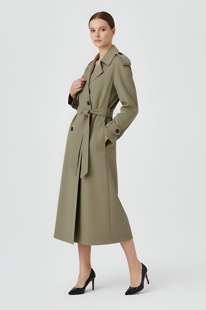 A woman dressed in a long green trench coat stands against a plain white background.