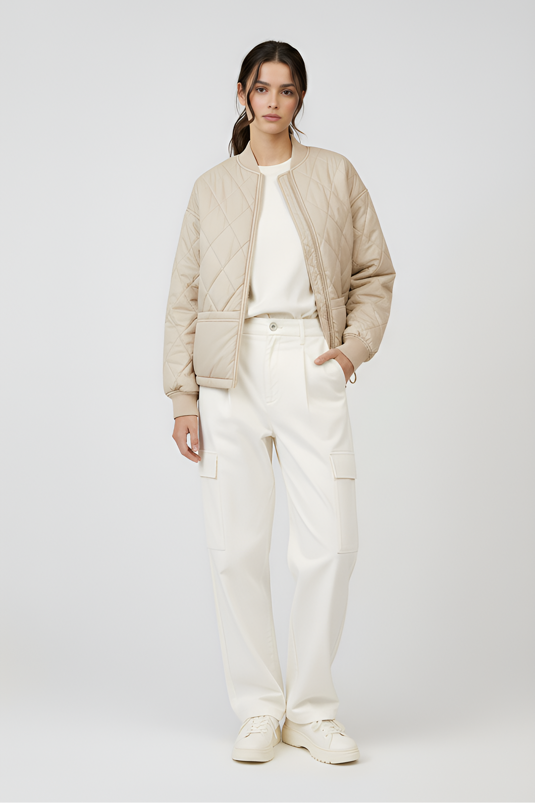 Woman wearing a beige bomber jacket and white pants on a light gray background