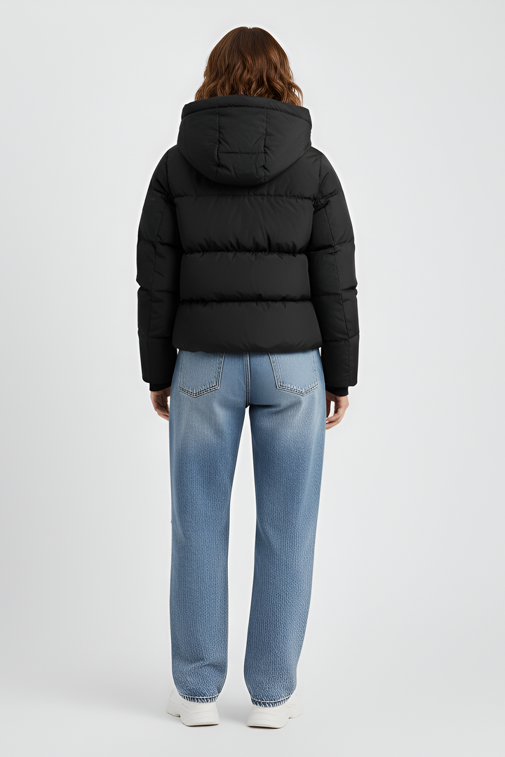 Woman wearing a black puffer jacket, blue jeans, and black boots on a plain background