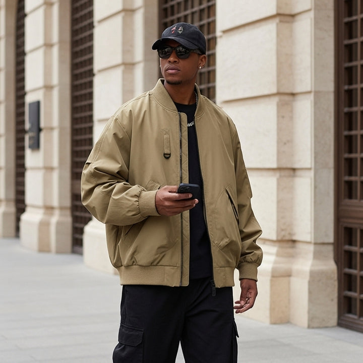 Man wearing a beige bomber jacket and black cap, standing in front of a building.