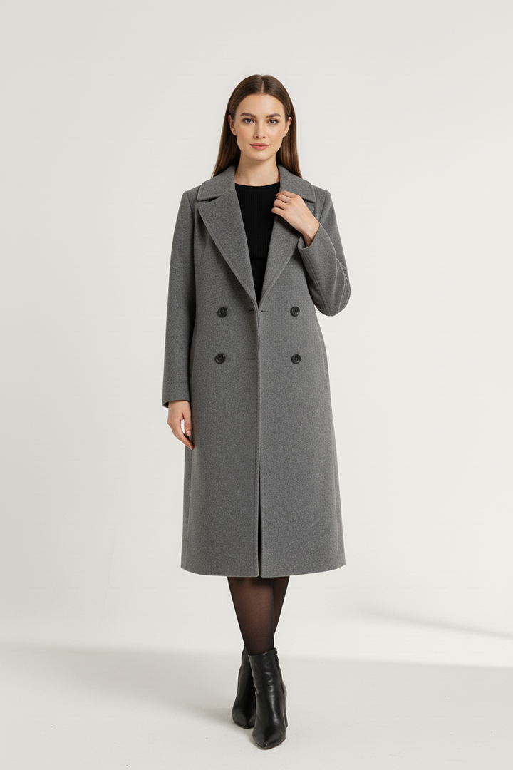 Mirella, a woman dressed in a gray coat, stands against a plain white background.