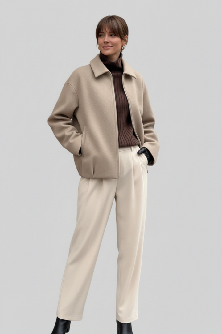 Person wearing a beige coat, brown sweater, and white pants on a light gray background