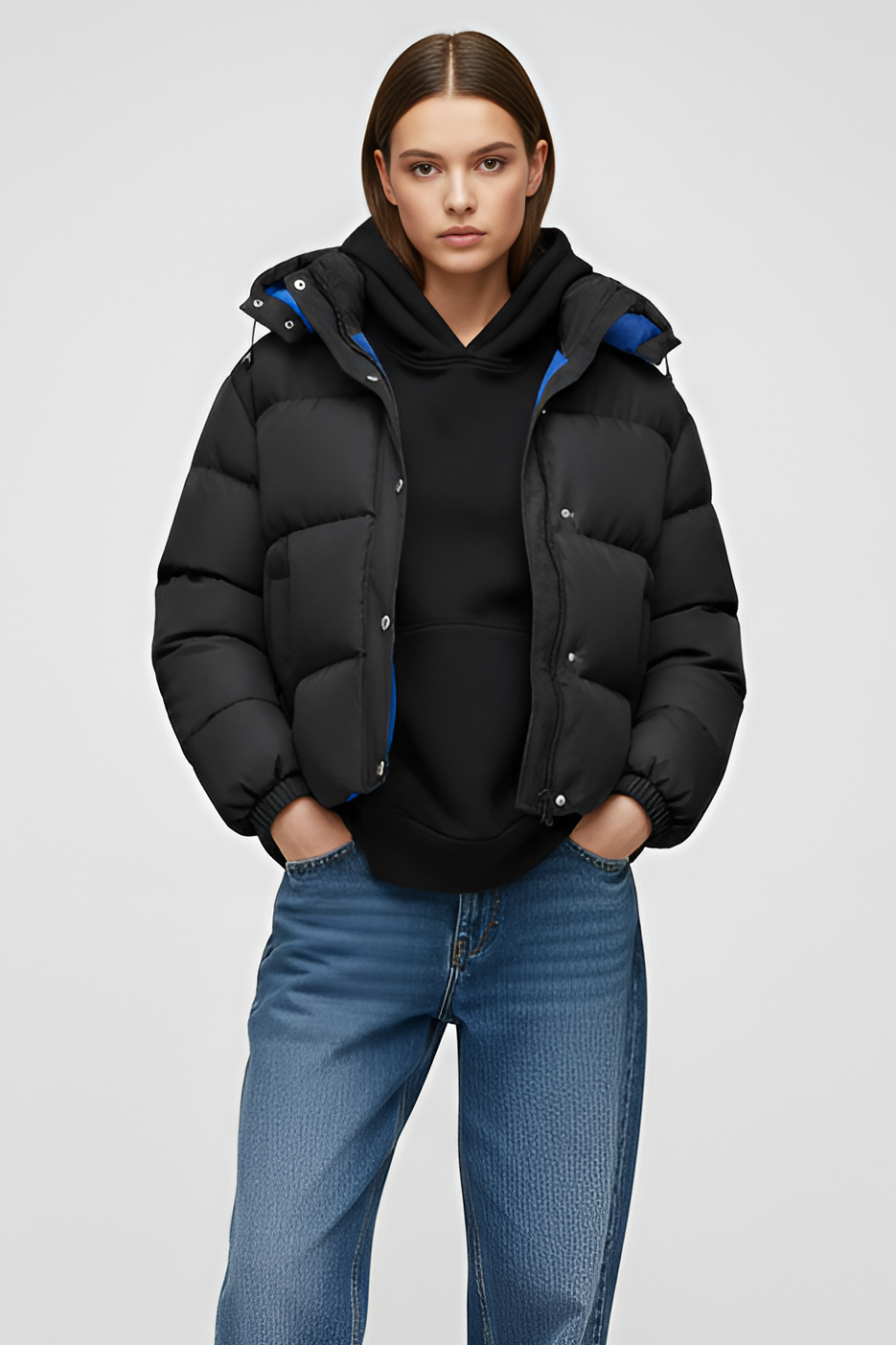 Person wearing a black puffer jacket, black hoodie, blue jeans, and white sneakers on a light gray background