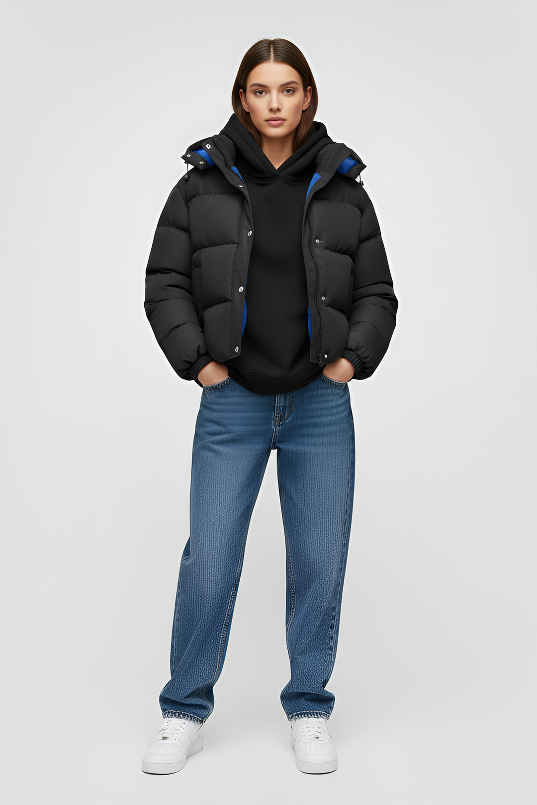 Person wearing a black puffer jacket, black hoodie, blue jeans, and white sneakers on a light gray background