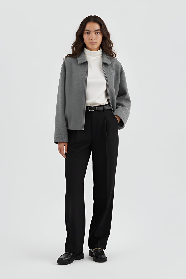 Person wearing a gray blazer, white turtleneck, black pants, and black shoes on a light gray background