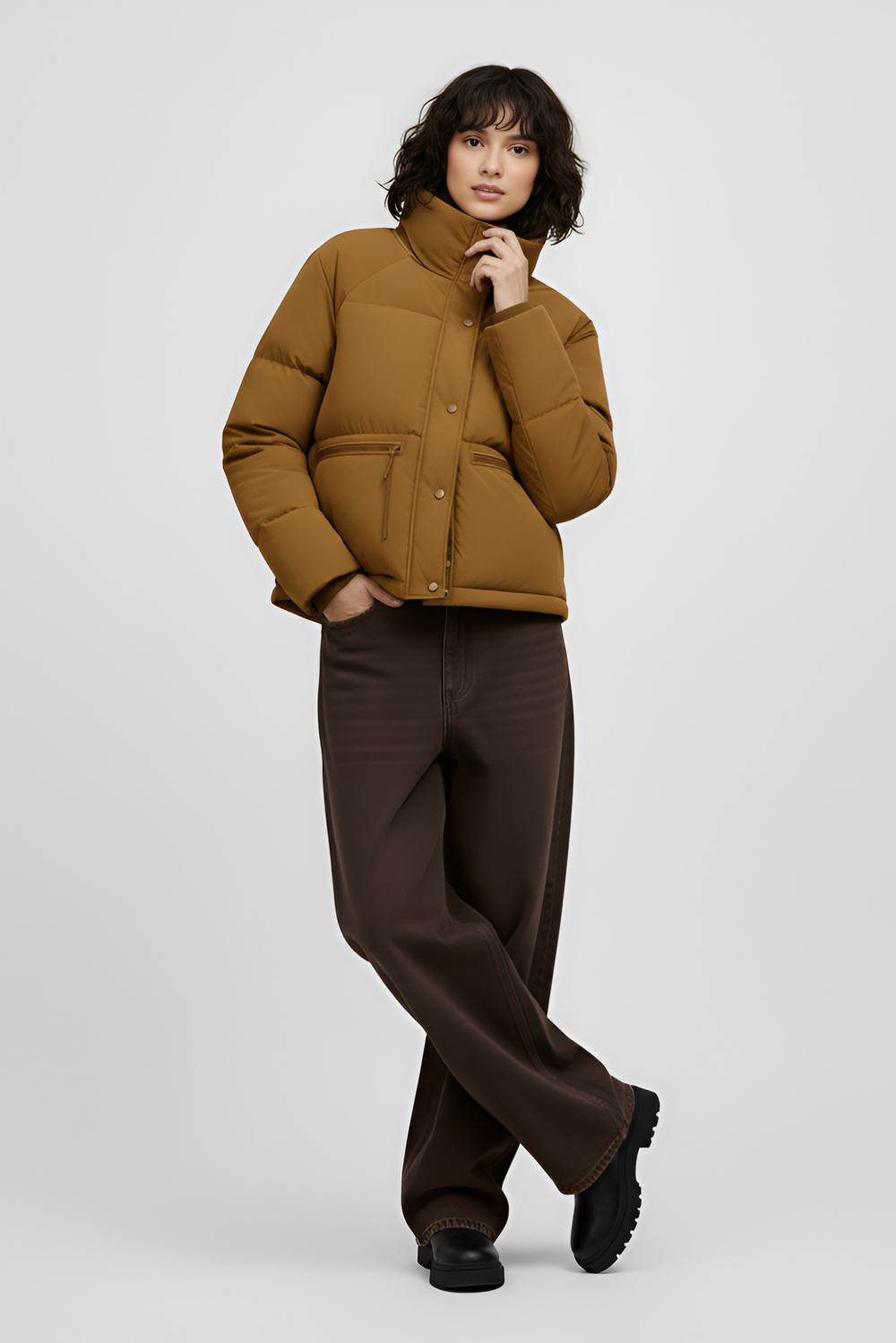 Woman wearing a Turmeric puffer jacket, brown sweater, black pants, and white shoes on a light gray background