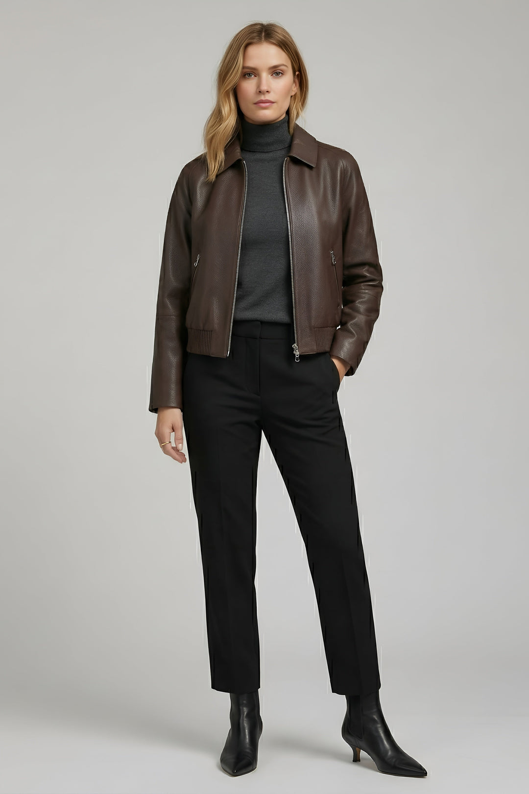 Woman wearing a brown leather jacket, black turtleneck, black pants, and black boots on a gray background