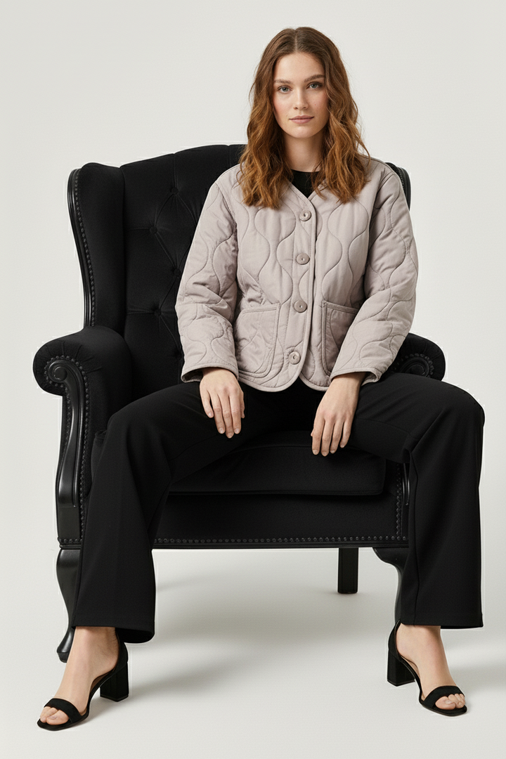 Woman sitting on a black armchair wearing a beige quilted jacket and black pants.