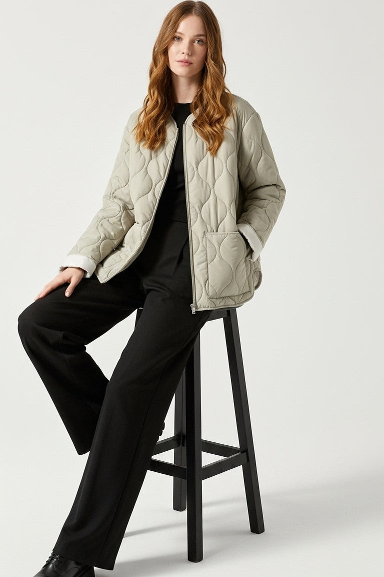 Woman sitting on a stool wearing a beige quilted jacket over a black outfit on a light gray background