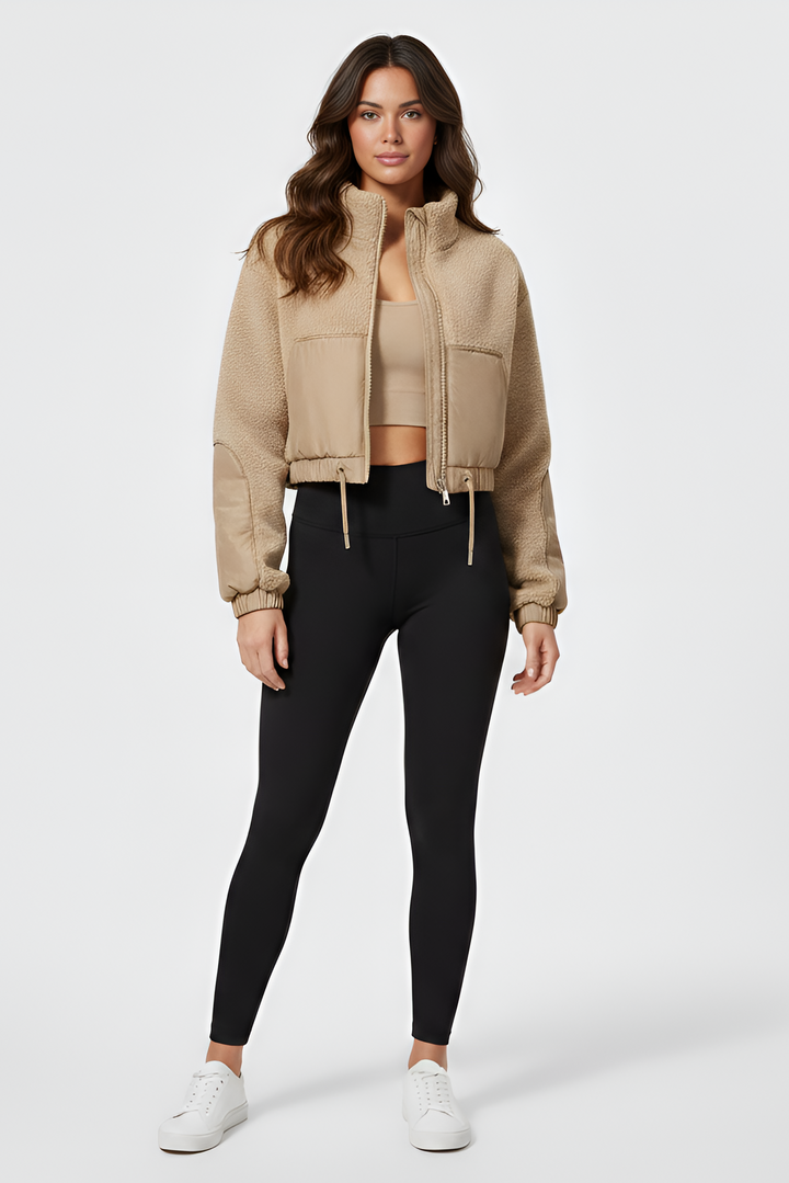 Woman wearing a beige cropped jacket, black leggings, and white sneakers on a white background