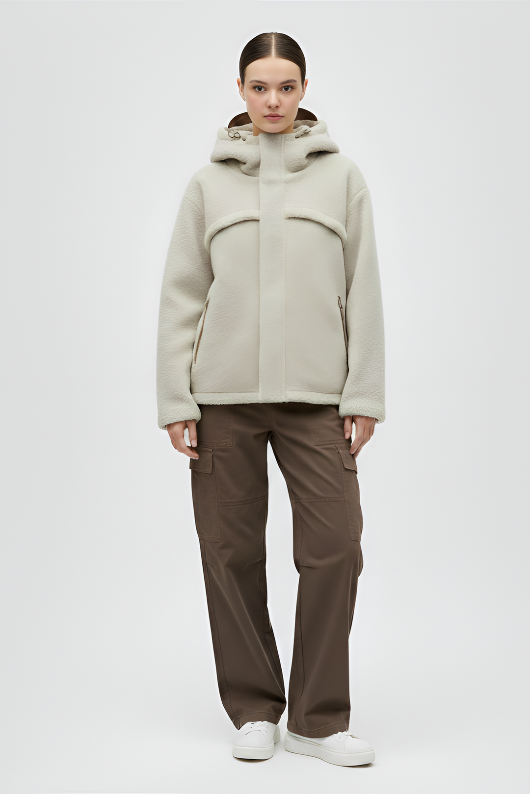 Woman wearing a beige faux fur jacket, white shirt, black cargo pants, and white sneakers on a gray background