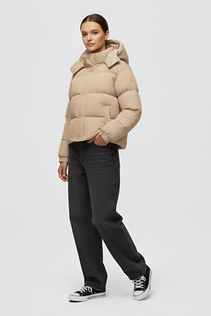 Woman wearing a beige puffer jacket and cap standing in an urban setting.