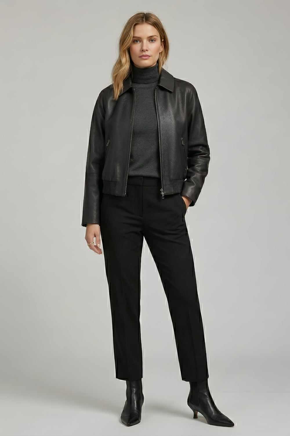 Woman wearing a black leather jacket, black turtleneck, black pants, and black boots on a gray background