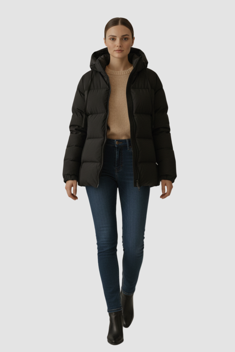 Woman wearing a black puffer jacket, beige sweater, blue jeans, and black boots on a neutral background