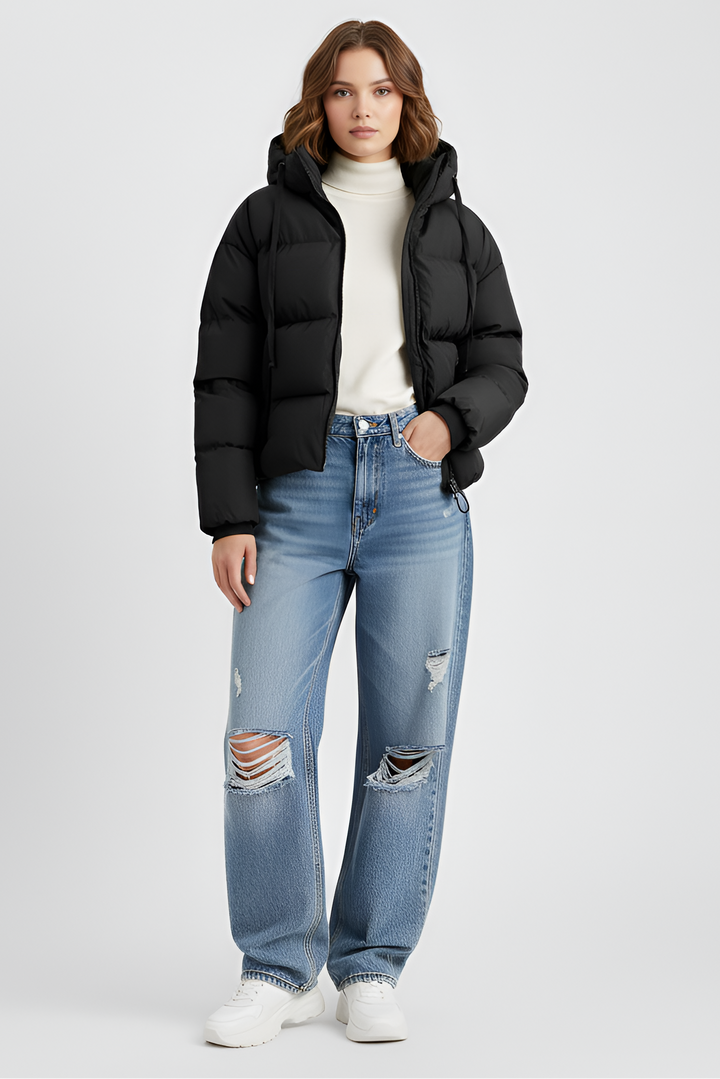 Woman wearing a black puffer jacket, blue jeans, and black boots on a plain background