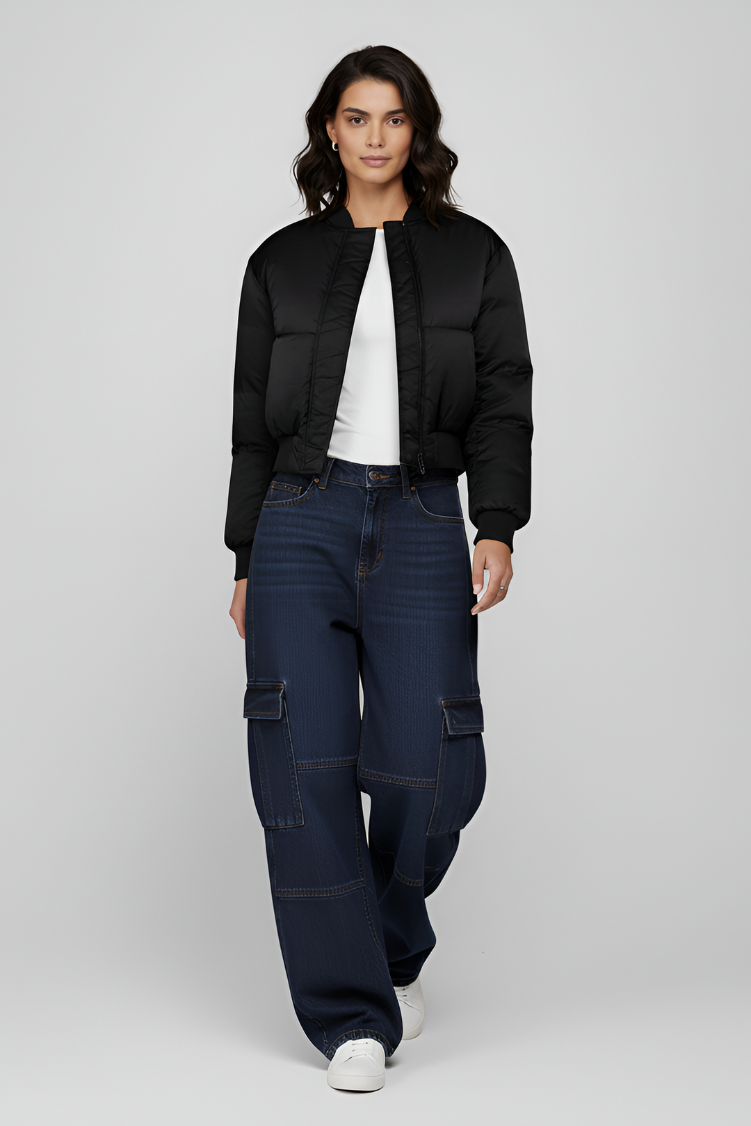Woman wearing a black puffer jacket, white shirt, and dark blue jeans on a light gray background