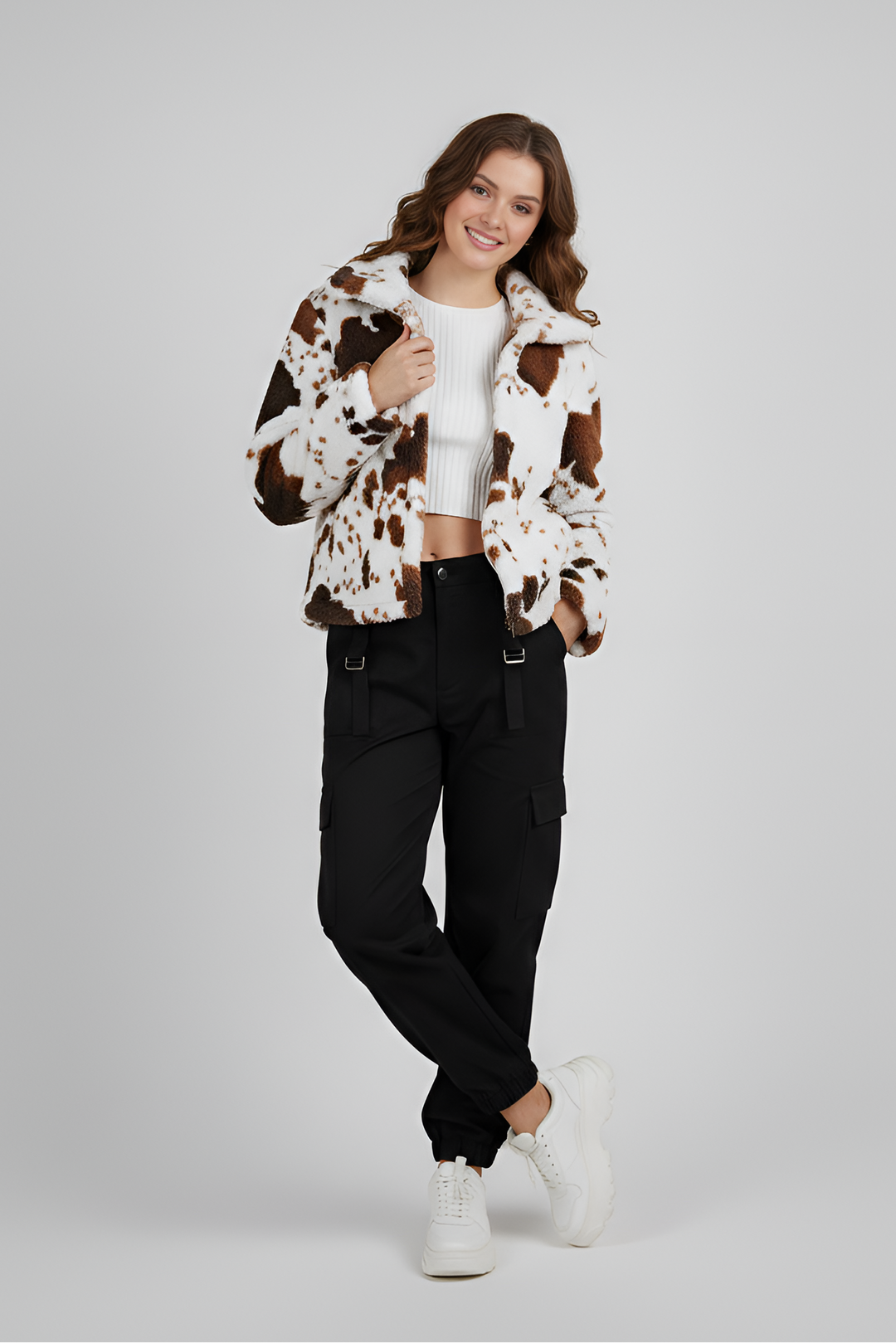 Woman wearing a cow print jacket, white top, black pants, and white sneakers on a gray background