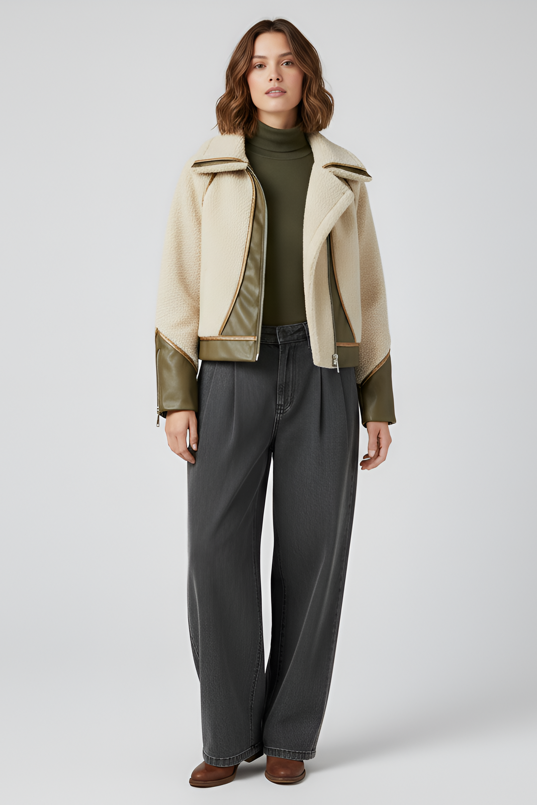 Woman wearing a cream fleece jacket with olive leather accents and wide dark gray trousers, standing against a light backdrop