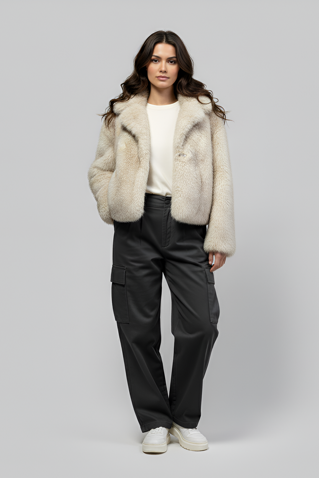 Woman wearing a cream fur coat, white sweater, black cargo pants, and white sneakers on a gray background