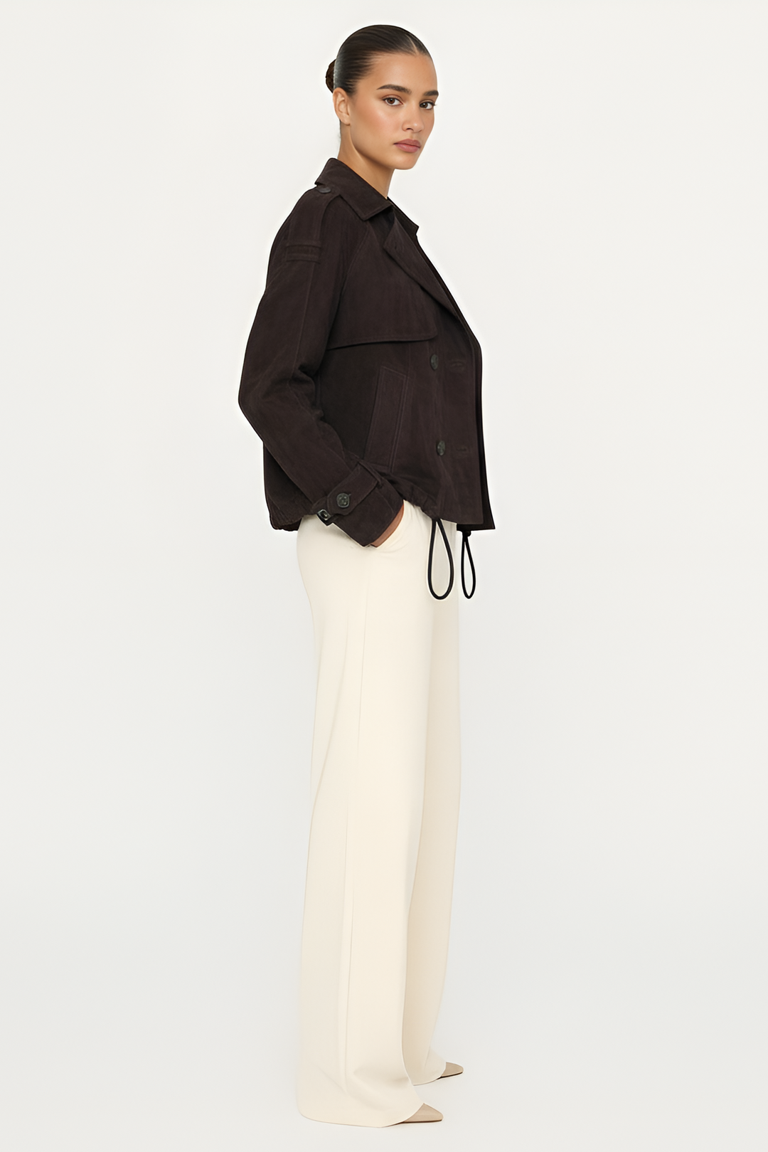 Woman wearing a dark brown jacket and white pants on a light gray background