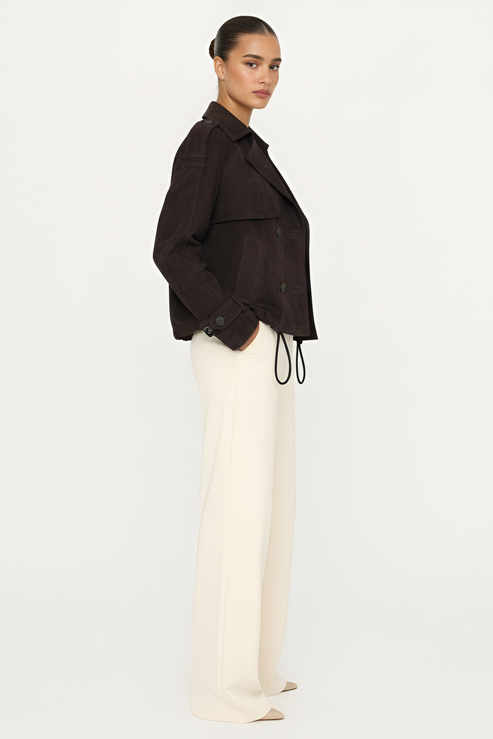 Woman wearing a dark brown jacket and white pants on a light gray background