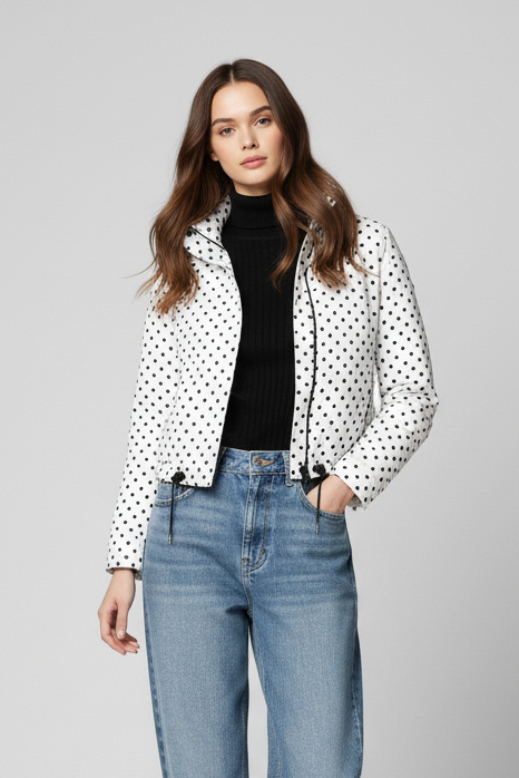 Woman wearing a polka dot jacket, black turtleneck, blue jeans, and white sneakers on a gray background