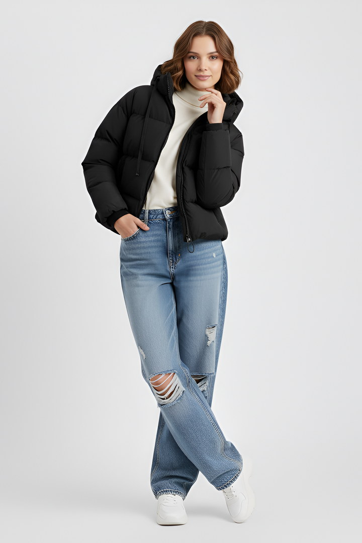 Woman wearing a black puffer jacket, blue jeans, and black boots on a plain background
